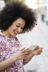 Portrait of smiling young woman with earphones looking at cell phone - KKAF00701