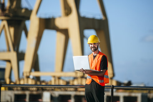 Porträt eines lächelnden Hafenarbeiters, der einen Laptop bei der Arbeit benutzt - KIJF01379