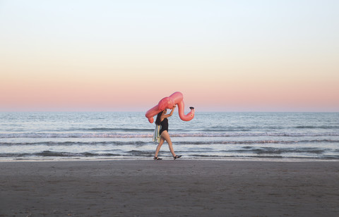 Young woman walking with inflatable pink flamingo on the beach at sunset stock photo