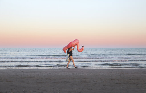 Junge Frau geht mit aufblasbarem rosa Flamingo am Strand bei Sonnenuntergang - RTBF00812