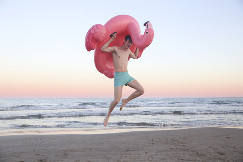 Lachender junger Mann, der am Strand mit einem aufblasbaren rosa Flamingo in die Luft springt, lizenzfreies Stockfoto