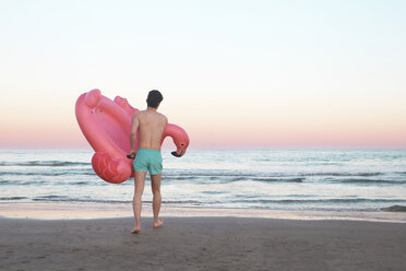 Back view of young man with inflatable pink flamingo on the beach - RTBF00816