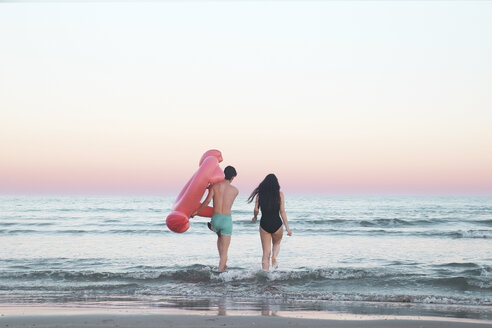 Rückenansicht eines jungen Paares, das mit einem aufblasbaren rosa Flamingo ins Meer geht - RTBF00817