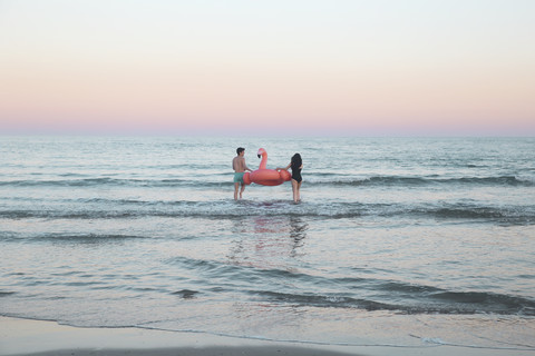 Rückenansicht eines jungen Paares, das mit einem aufblasbaren rosa Flamingo ins Meer geht, lizenzfreies Stockfoto