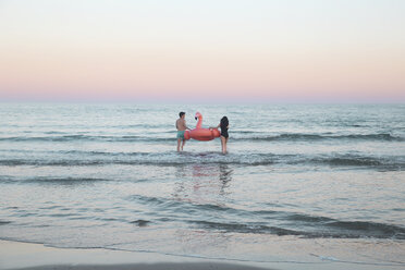 Rückenansicht eines jungen Paares, das mit einem aufblasbaren rosa Flamingo ins Meer geht - RTBF00818