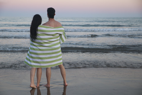 Rückenansicht des jungen Paares am Strand zusammen in Handtuch gewickelt beobachten Sonnenuntergang, lizenzfreies Stockfoto