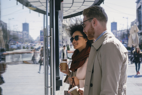 Pärchen in der Stadt mit Kaffee zum Mitnehmen, das in ein Schaufenster schaut, lizenzfreies Stockfoto