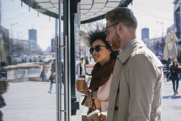 Pärchen in der Stadt mit Kaffee zum Mitnehmen, das in ein Schaufenster schaut - MOMF00142