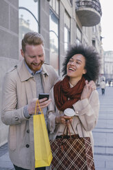 Glückliches Paar in der Stadt mit Handy und Einkaufstaschen - MOMF00151