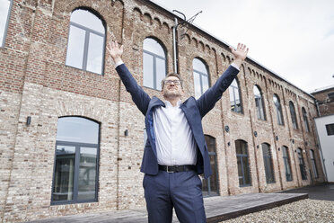 Businessman standing in front of building with arms raised - FMKF03863