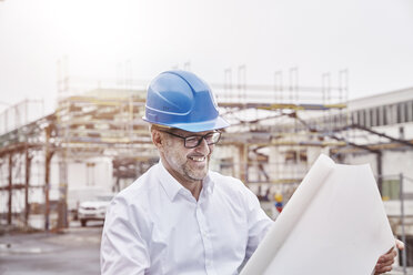 Portrait of laughing man wearing blue hart hat looking at construction plan - FMKF03864