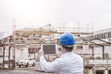 Back view of man wearing blue hart hat taking picture with tablet at construction site - FMKF03868