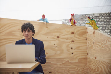 Junger Geschäftsmann arbeitet am Laptop mit Handpuppen, die ihn von einer Holzwand aus beobachten - RHF01874