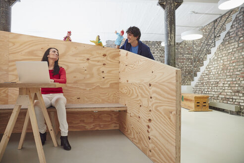 Businesswoman working on laptop with colleagues playing hand puppets behind wooden wall - RHF01876