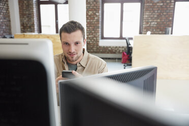 Junger Mann sitzt im Büro und benutzt Computer und Smartphone - RHF01918