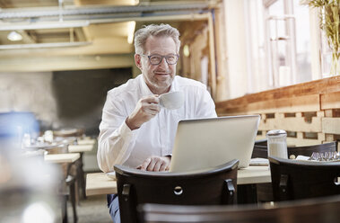 Portrait of confident mature businessman in cafe using laptop - FMKF03914