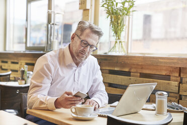 Mature businessman in cafe using laptop and cell phone - FMKF03942