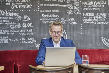 Portrait of confident mature businessman in cafe using laptop - FMKF03952