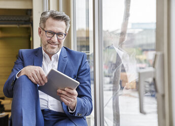 Smiling mature businessman using tablet at the window - FMKF03961