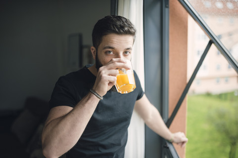 Junger Mann trinkt einen Orangensaft am Fenster, lizenzfreies Stockfoto