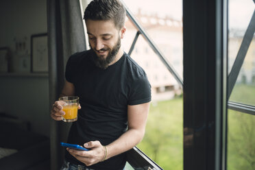 Junger Mann trinkt einen Orangensaft und benutzt sein Smartphone am Fenster - RAEF01832