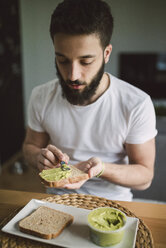 Junger Mann beim Frühstück zu Hause, Toast mit Avocadocreme - RAEF01837