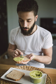 Junger Mann beim Frühstück zu Hause, Toast mit Avocadocreme - RAEF01837