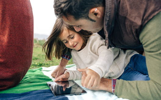 Glückliches Mädchen und ihr Vater benutzen ein Tablet und liegen auf einer Decke im Freien - DAPF00704