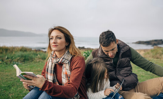 Relaxed woman with book looking aside while girl and man using tablet outdoors - DAPF00705