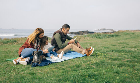 Family with dog sitting on blanket at the coast using wireless devices - DAPF00707