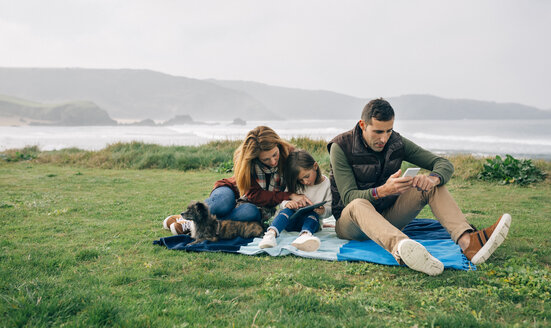 Familie mit Hund sitzt auf einer Decke an der Küste und benutzt drahtlose Geräte - DAPF00708