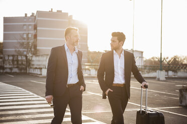 Two businessmen with rolling suitcase on parking place at sunset - DIGF01978