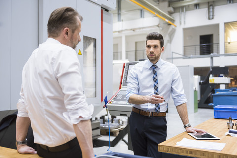 Zwei Geschäftsleute an einem Tisch in einer Fabrikhalle, die sich unterhalten, lizenzfreies Stockfoto