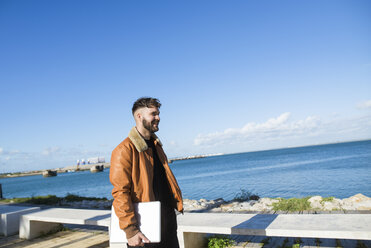 Spanien, Cadiz, Junger Mann am Hafen, der mit einem Laptop unterwegs ist - KIJF01391