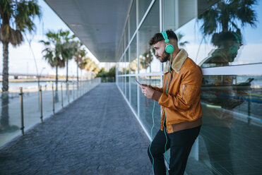 Spain, Cadiz, Young man at the harbour using smartphone, wearing headphones - KIJF01397
