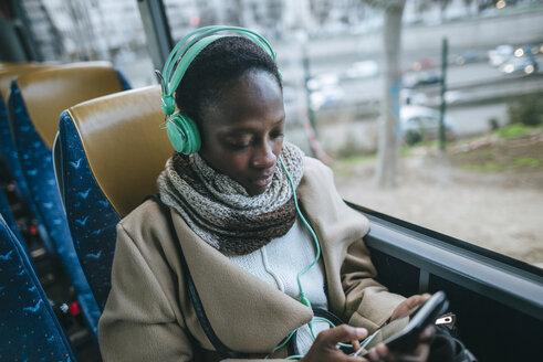 Junge Frau in einem Bus hört Musik mit Kopfhörern, während sie auf ihr Handy schaut - KIJF01412