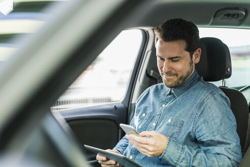 Businessman sitting in car using smartphone and digital tablet - UUF10347