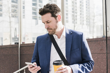 Businessman in the city, reading smart phone messages and holding cup of coffee - UUF10391