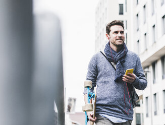 Businessman carrying skateboard, using smartphone and earphones - UUF10396