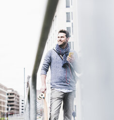 Businessman carrying skateboard, using smartphone and earphones - UUF10398