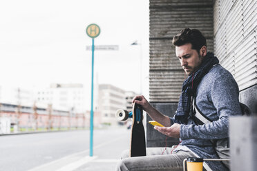 Businessman with skateboard sitting at bus stop using smartphone - UUF10399