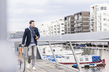 Businessman pushing bicycle in the city, while using smartphone and earphones - UUF10403