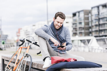 Geschäftsmann in der Stadt mit Fahrrad, Smartphone und Kopfhörer - UUF10407