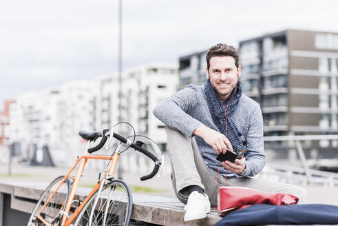 Geschäftsmann in der Stadt mit Fahrrad, Smartphone und Kopfhörer - UUF10408