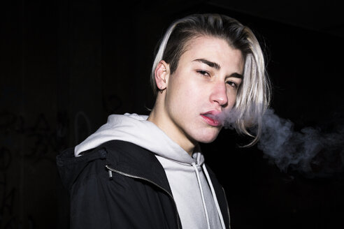 Portrait of young man exhaling cigarette smoke in front of black background - ABZF01959