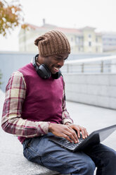 Lächelnder Mann mit Kopfhörern, der auf einer Bank sitzt und einen Laptop benutzt - MAUF01044
