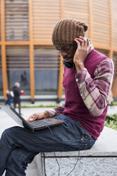 Mann sitzt auf einer Bank, benutzt Kopfhörer und schaut auf einen Laptop - MAUF01046