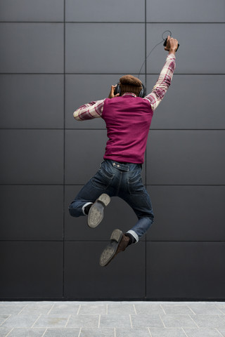 Rückansicht eines glücklichen Mannes, der in die Luft springt, während er mit Kopfhörern und Mobiltelefon Musik hört, lizenzfreies Stockfoto