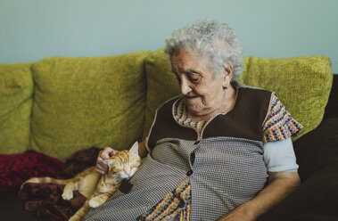 Ältere Frau sitzt auf der Couch und streichelt eine getigerte Katze - RAEF01860