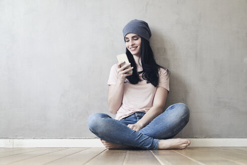 Lächelnde junge Frau, die auf dem Boden sitzt und ein Mobiltelefon benutzt - FMKF03991